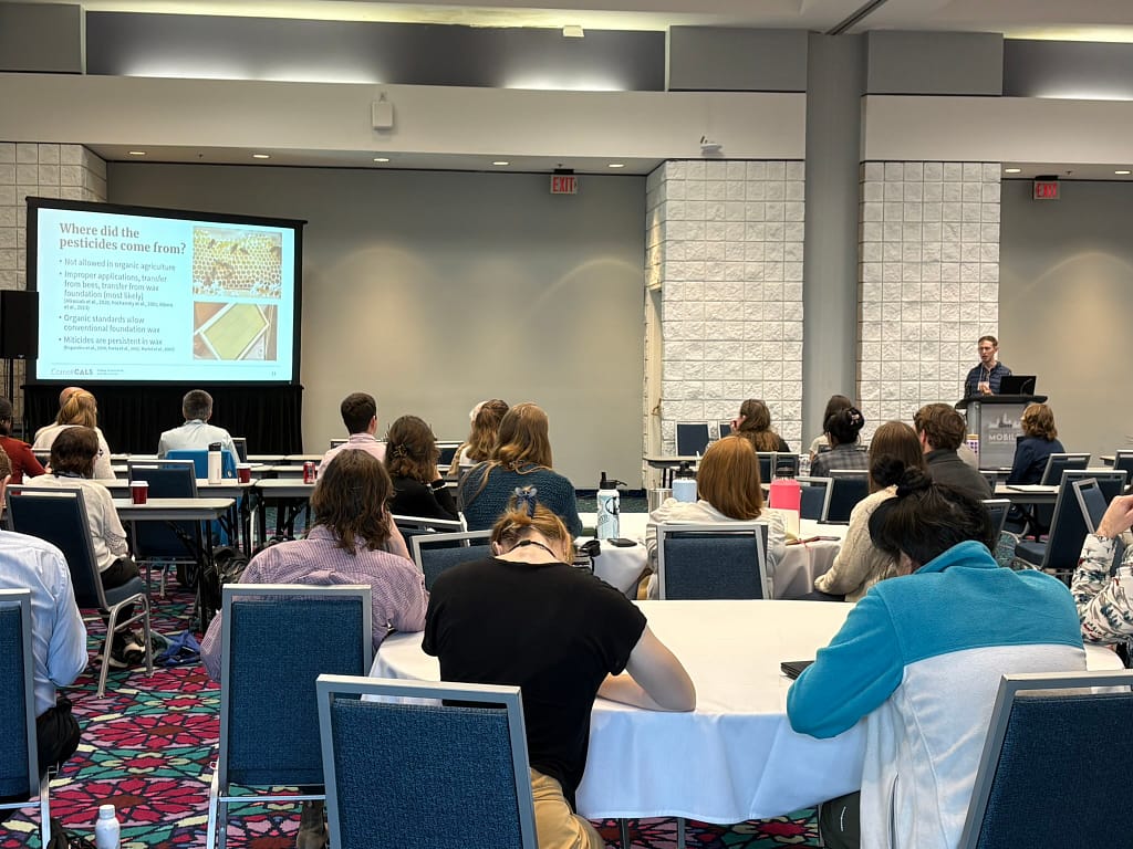 A researcher at the front of a room delivers a slideshow presentation to a room of conference attendees seated at several round tables