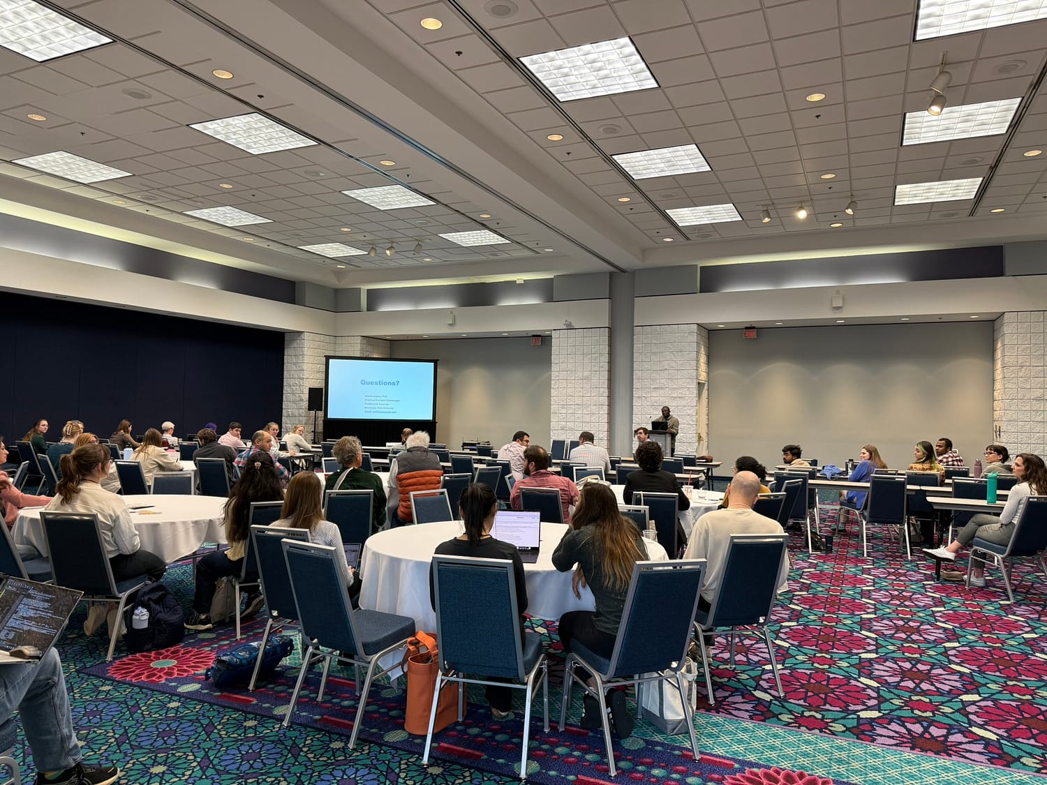 A researcher at the front of a room delivers a slideshow presentation to a room of conference attendees seated at several round tables